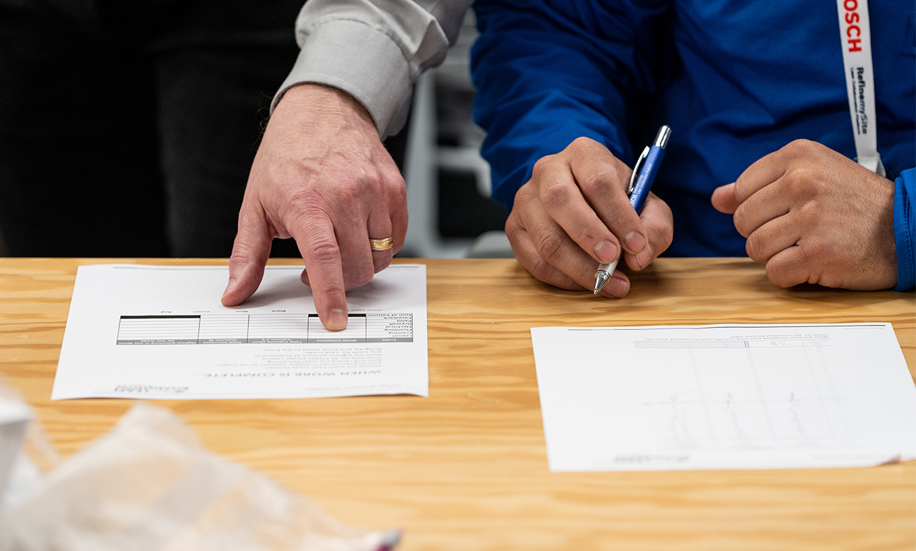 2 men's hands with one holding a pen and another pointing at 2 sheets of paper