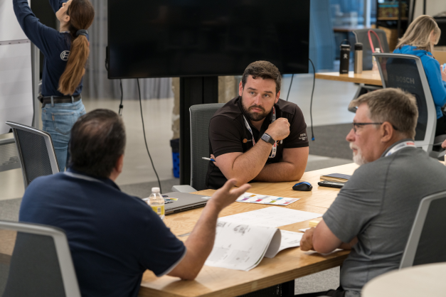 Three men sit at a meeting table and discuss Lean methods and services. In the background, a woman hooks up a TV monitor