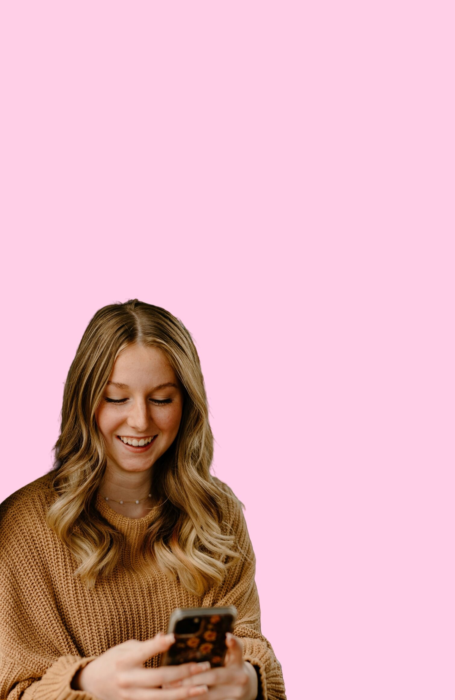 A young woman smiling while looking at her phone, set against a pink background.
