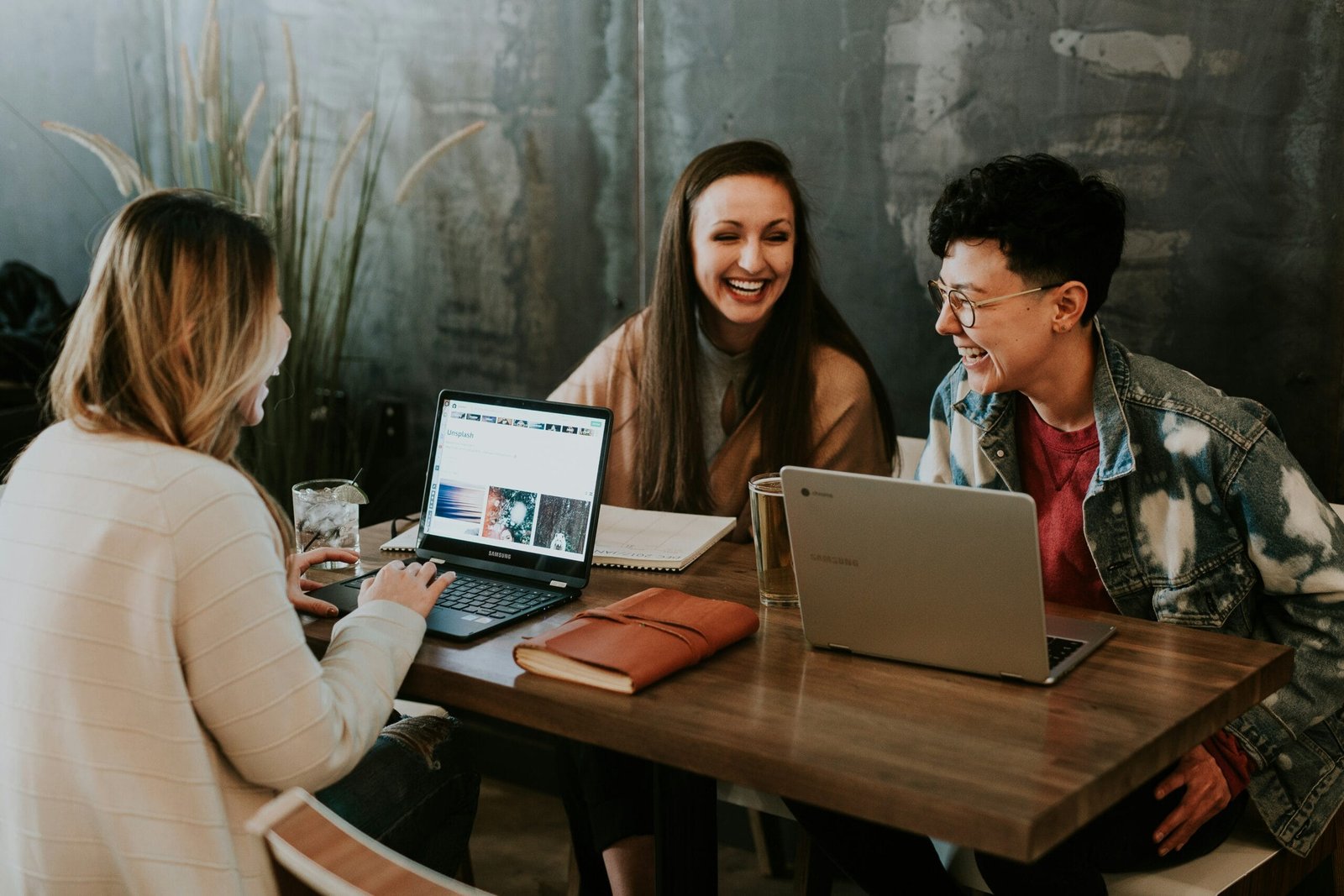 People working together on a desk. Laptops are on the table. They are all smiling and looking happy. The table is wooden with an industrial wall behind it.