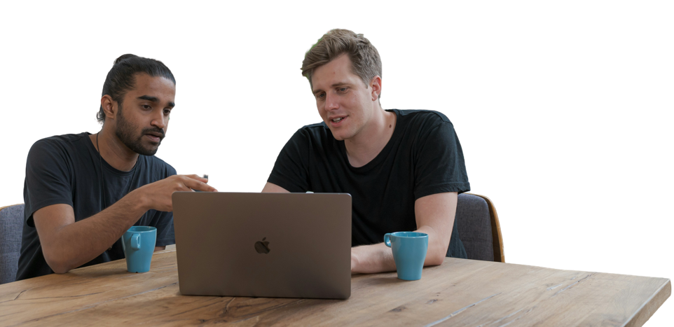 Two men, one mixed race and the other a white man looking at a computer drinking coffee on a table.